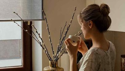 A woman sitting by a window holding a cup, looking reflective in a softly lit living space, capturing a quiet moment that inspires the reminder to be grateful for the ordinary.